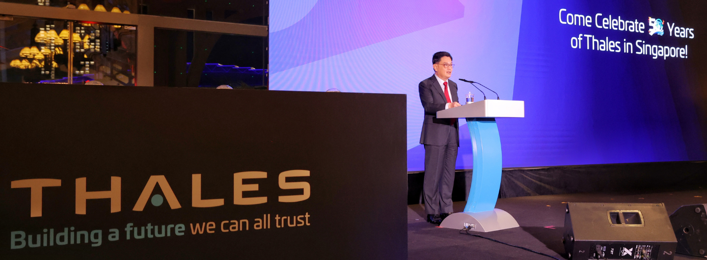 Man at podium on stage; "THALES" logo visible, screen reads "Come Celebrate 50 Years of Thales in Singapore!".